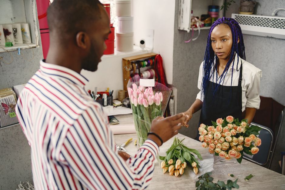 A florist with vibrant purple braids is wrapping a bouquet of pink tulips in light pink floral paper at Lovemeflowers.co.uk. The arrangement includes fresh, delicate tulips with green stems and leaves