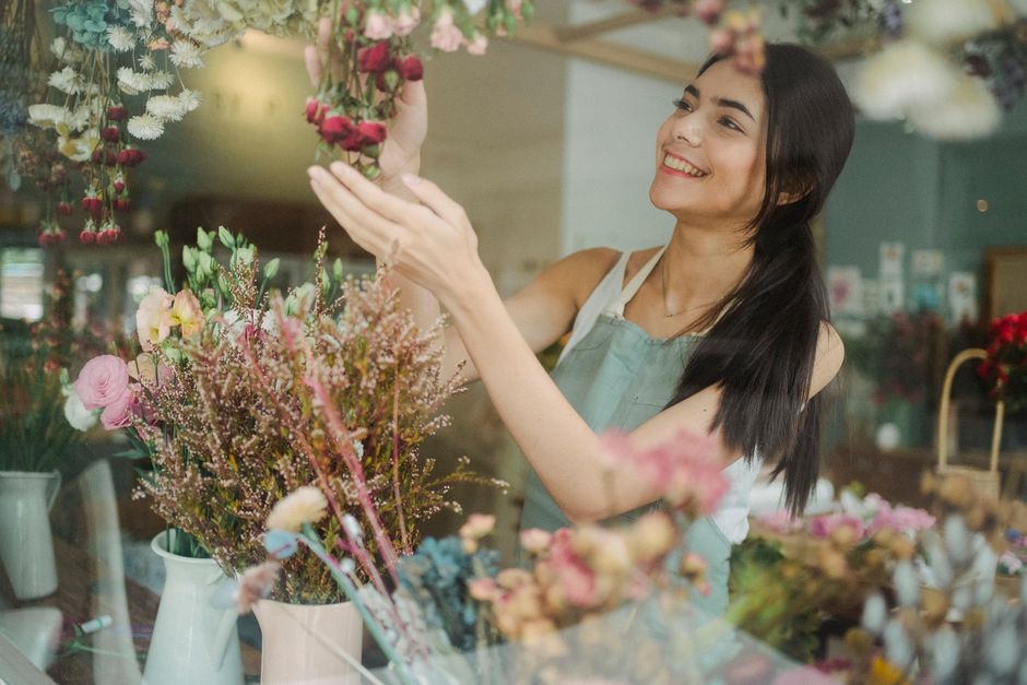A smiling young woman with dark hair tied back, wearing a light grey apron, is standing inside a flower shop holding a white ceramic pitcher filled with fresh pink carnations and white lisianthus. The