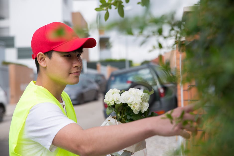 A young male floral delivery courier wearing a red cap and yellow high-visibility vest is seen outdoors, carefully placing a bouquet of white hydrangeas wrapped in light-colored paper onto a doorstep
