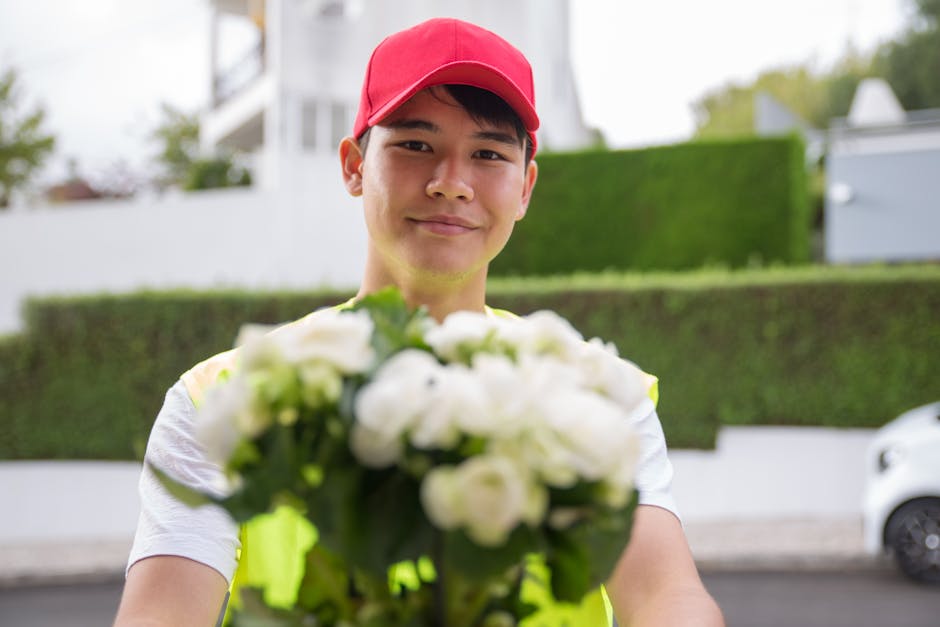 A young male floral delivery courier wearing a red cap and yellow high-visibility vest is seen outdoors, carefully placing a bouquet of white hydrangeas wrapped in light-colored paper onto a doorstep