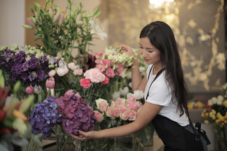 A woman with light brown hair and bangs, wearing a beige sweater, is smiling while talking on her mobile phone and holding a large, vibrant bouquet of fresh flowers. The bouquet features a variety of