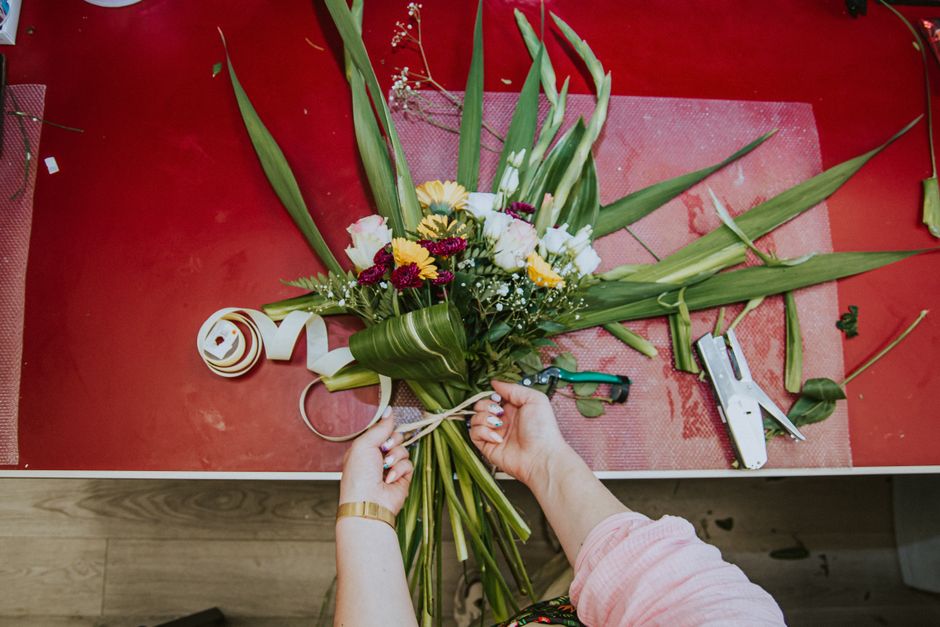 A rustic wooden table covered with brown craft paper, displaying various fresh flowers, green foliage, and floral arrangement tools. Visible are white daisies with pink centers, yellow blooms, and del