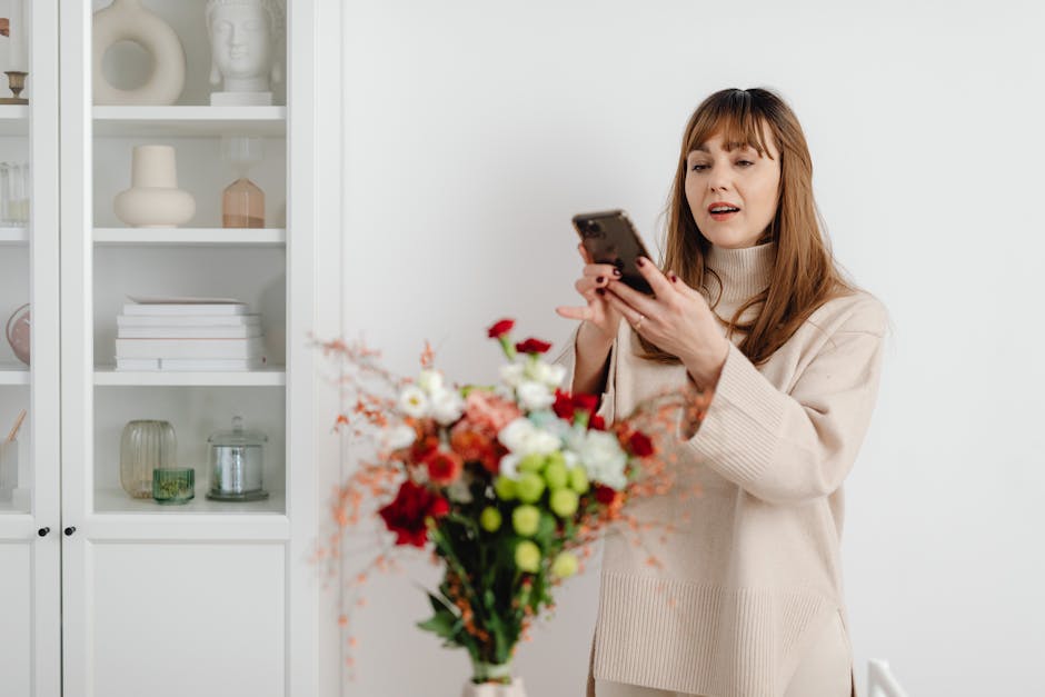 A female florist and a customer are engaged in a floral arrangement at a flower shop, surrounded by a variety of fresh flowers including pink peonies, white lilies, and red carnations. The florist, dr