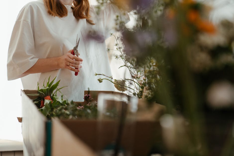 Close-up of a person using pruning shears to trim a small branch, with a floral arrangement on a red surface in the foreground. The arrangement includes various stems and greenery, some with visible l