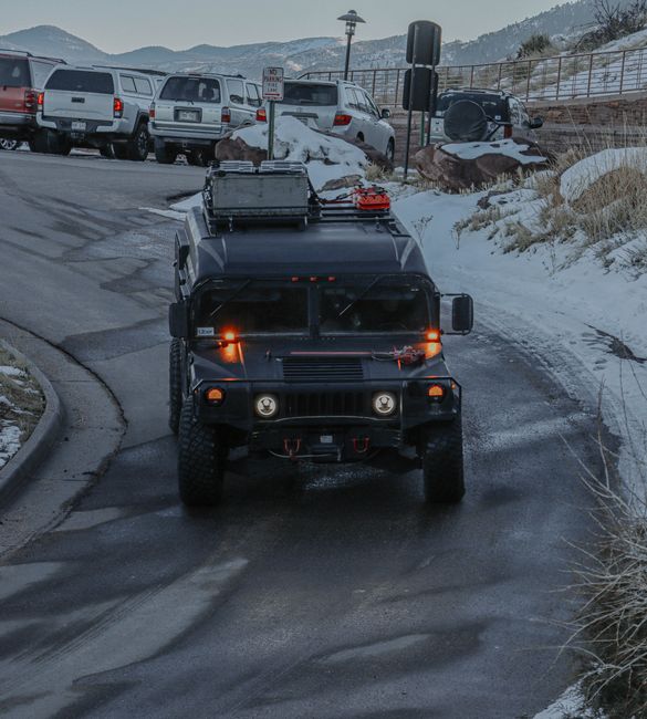 A grey military-style vehicle is positioned at a petrol station with multiple fuel pumps, situated outdoors under a large canopy. Surrounding the vehicle are modern cars parked nearby, and the backgro