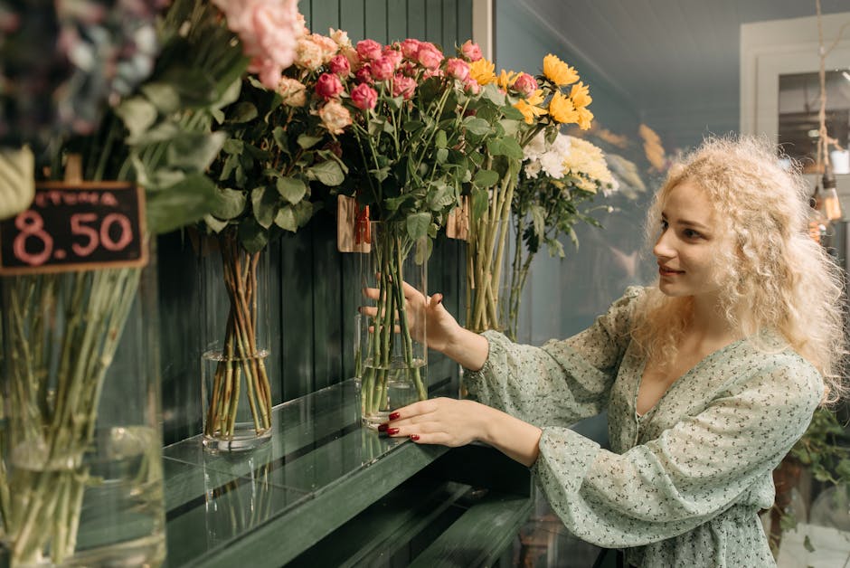 A young woman with curly blonde hair and fair skin selecting a bouquet of fresh flowers in a florist shop. She is wearing a light green floral-patterned dress with long sleeves and is smiling gently a