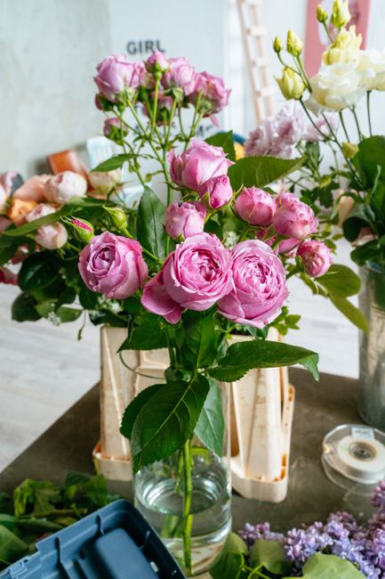 A young woman with curly blonde hair and fair skin selecting a bouquet of fresh flowers in a florist shop. She is wearing a light green floral-patterned dress with long sleeves and is smiling gently a