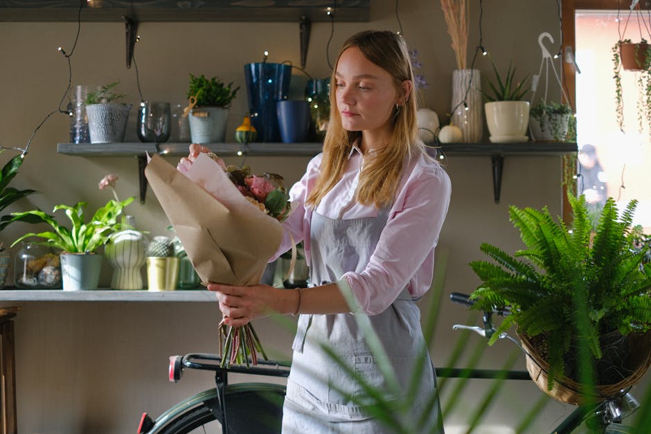 A young woman with long blonde hair, wearing a light pink shirt and a grey apron, is holding a bouquet of fresh flowers wrapped in light brown paper. The bouquet features pink roses, with some greener