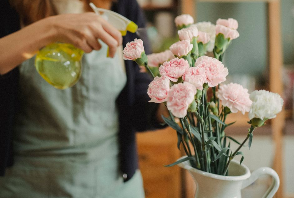 A young woman with long blonde hair, wearing a light pink shirt and a grey apron, is holding a bouquet of fresh flowers wrapped in light brown paper. The bouquet features pink roses, with some greener